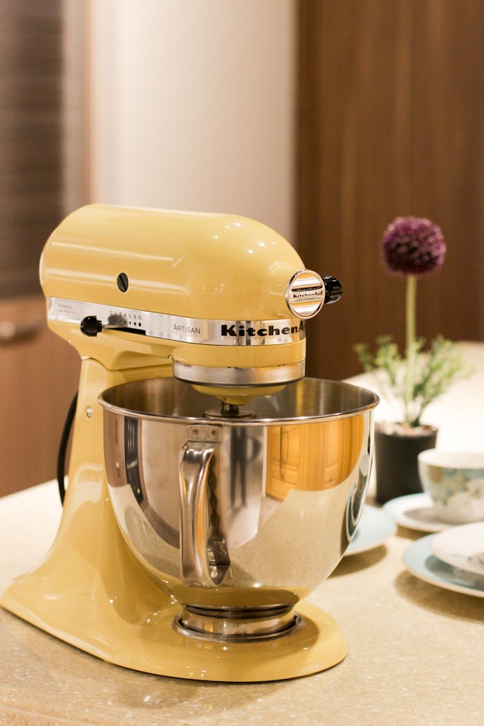 about-us-01 Vibrant yellow stand mixer on a kitchen countertop beside tableware and decor.