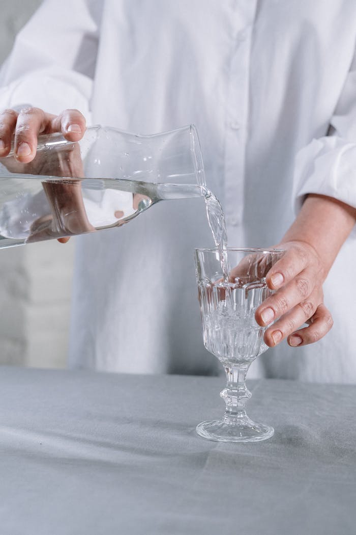 services-03 Close-up of hands pouring water from a jug into a crystal glass on a table.